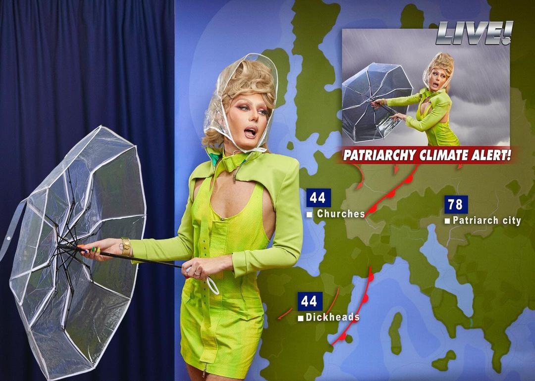A person in a green dress and clear rain gear stands with a clear umbrella against a weather map backdrop in a brightly lit photo studio.