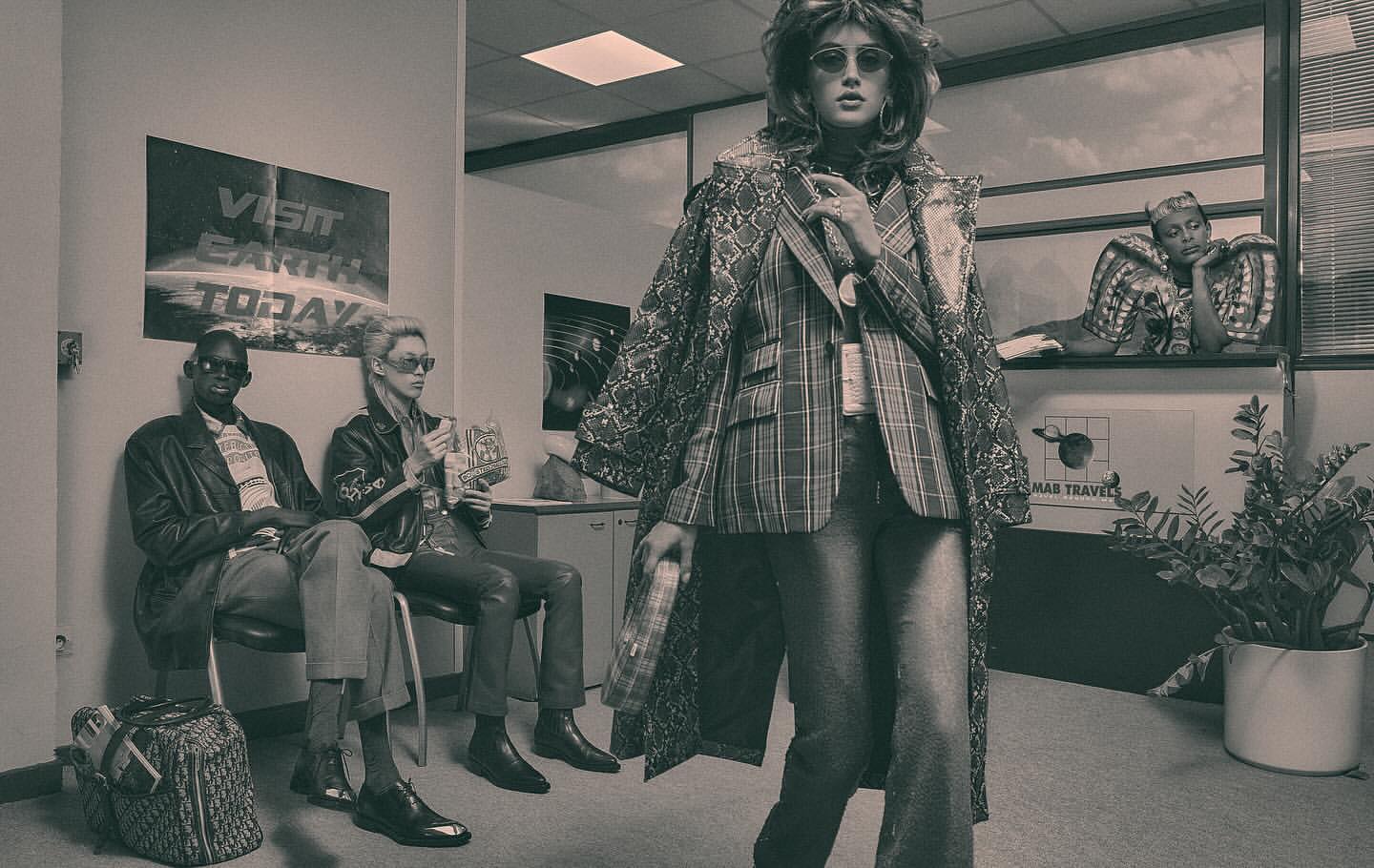 Monochromatic fashion photo featuring a woman in a plaid suit and snakeskin coat standing in a waiting room with seated and reclining figures.