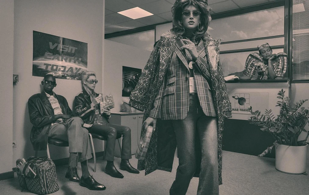 Monochromatic fashion photo featuring a woman in a plaid suit and snakeskin coat standing in a waiting room with seated and reclining figures.