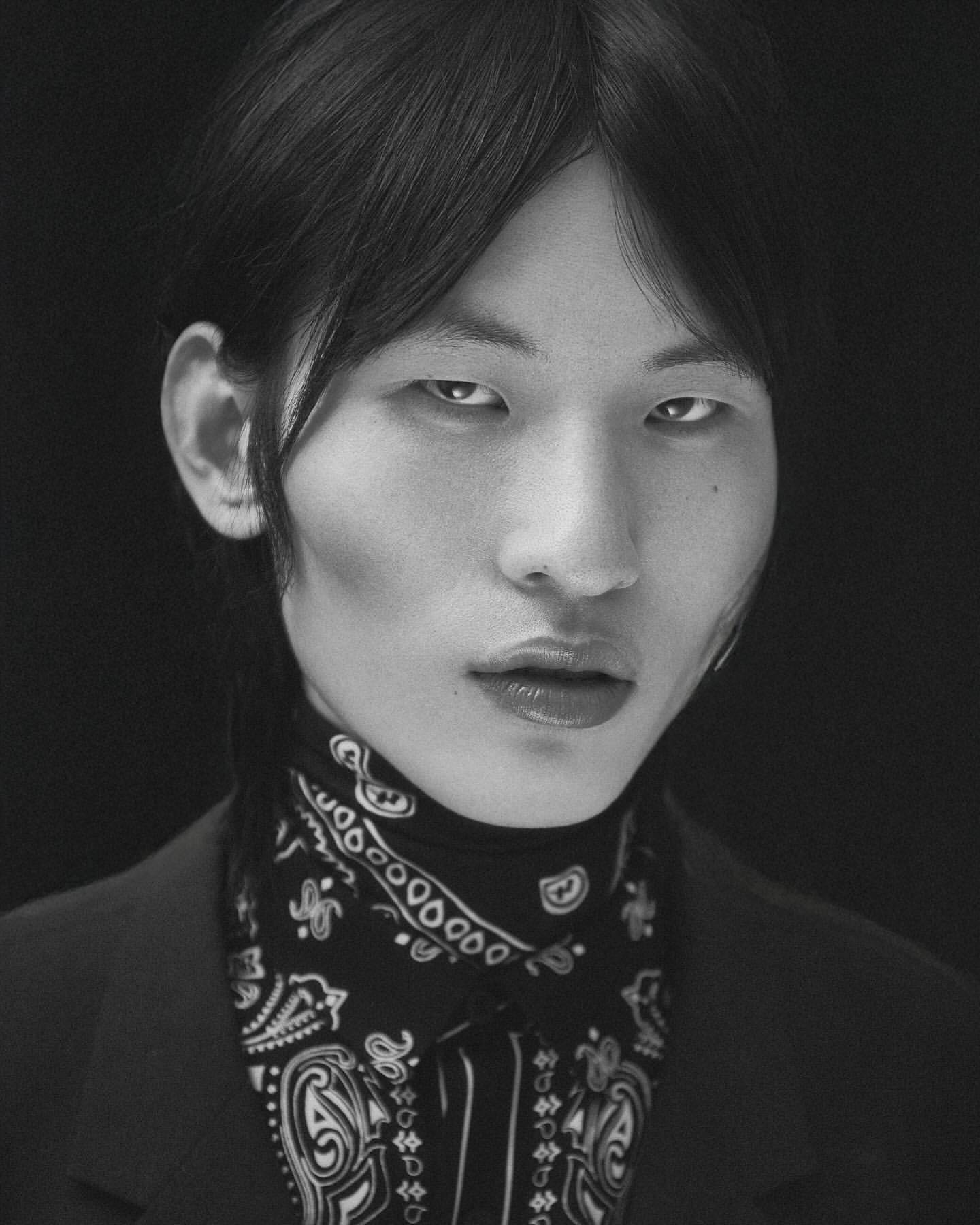 Striking black-and-white portrait of a person with dark hair, wearing a patterned bandana and dark suit jacket against a black backdrop.