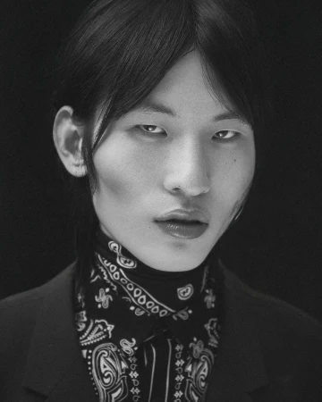 Striking black-and-white portrait of a person with dark hair, wearing a patterned bandana and dark suit jacket against a black backdrop.