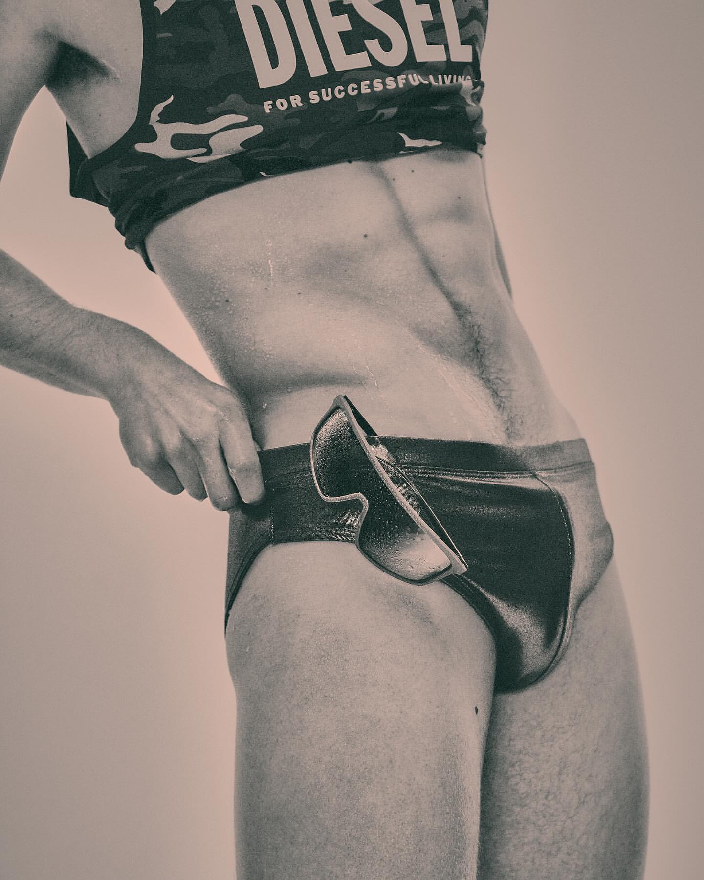 Man in briefs and tank top poses indoors. The tank top has the word DIESEL on it. He is touching the top of his briefs.