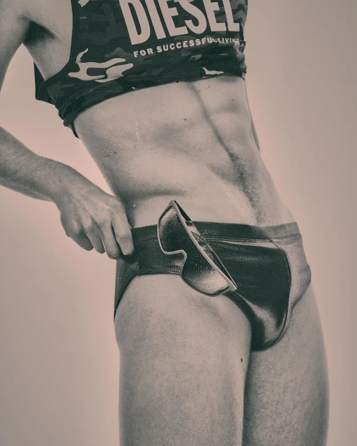 Man in briefs and tank top poses indoors. The tank top has the word DIESEL on it. He is touching the top of his briefs.
