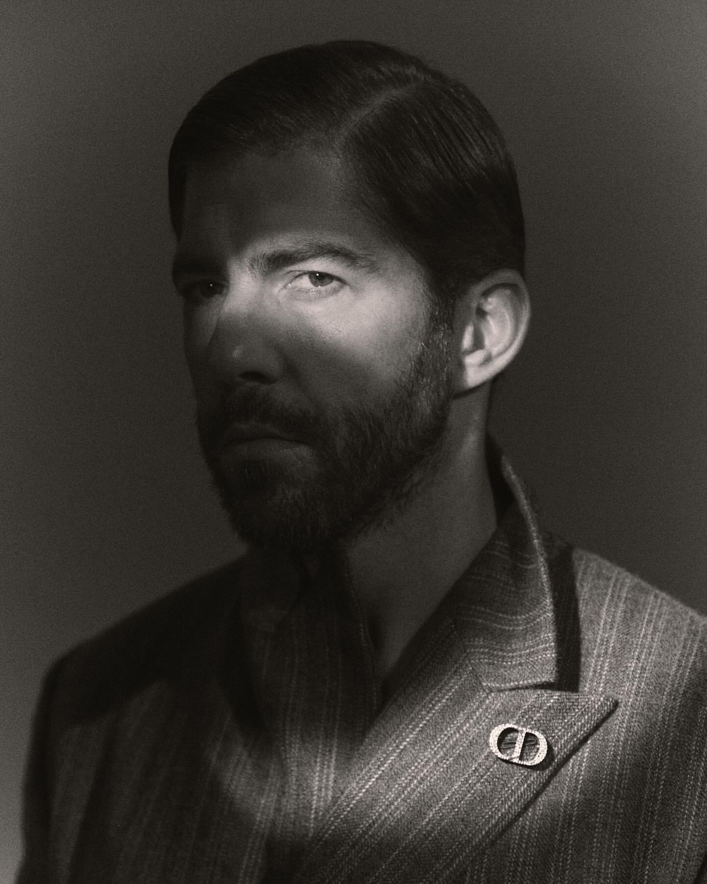 Black and white portrait of a handsome man with a beard wearing a striped suit with a designer lapel pin in a photo studio.