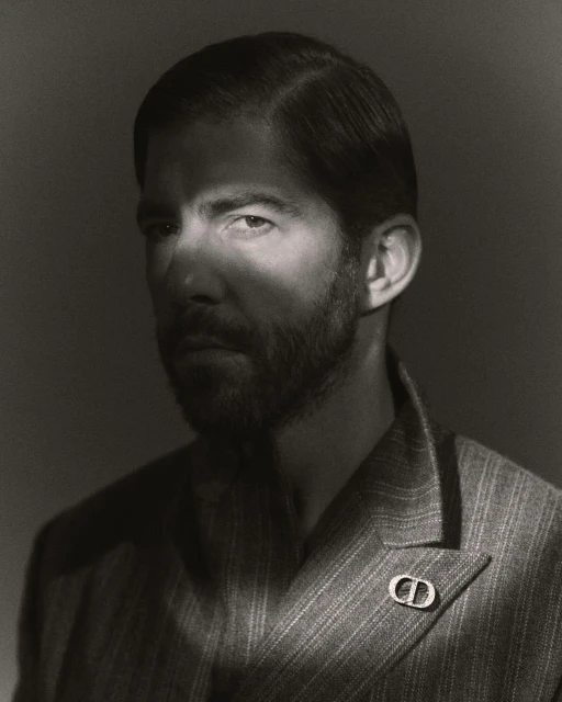 Black and white portrait of a handsome man with a beard wearing a striped suit with a designer lapel pin in a photo studio.