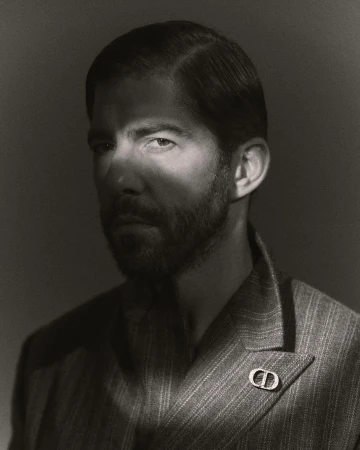 Black and white portrait of a handsome man with a beard wearing a striped suit with a designer lapel pin in a photo studio.