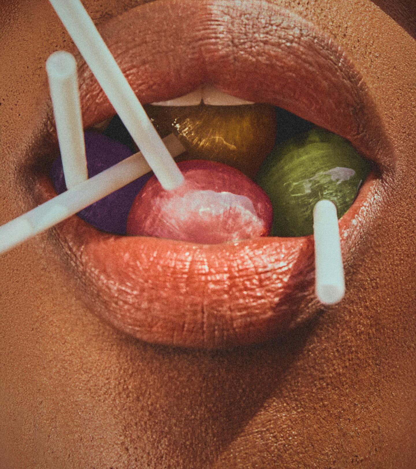 Close-up shot of a woman's mouth holding colorful lollipops with white sticks, lips painted with orange lipstick.