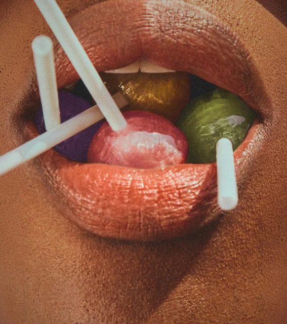 Close-up shot of a woman's mouth holding colorful lollipops with white sticks, lips painted with orange lipstick.