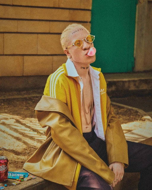 Portrait of a blonde adult with a bubble gum and sunglasses wearing a yellow track jacket and leaning against a wall outdoors.