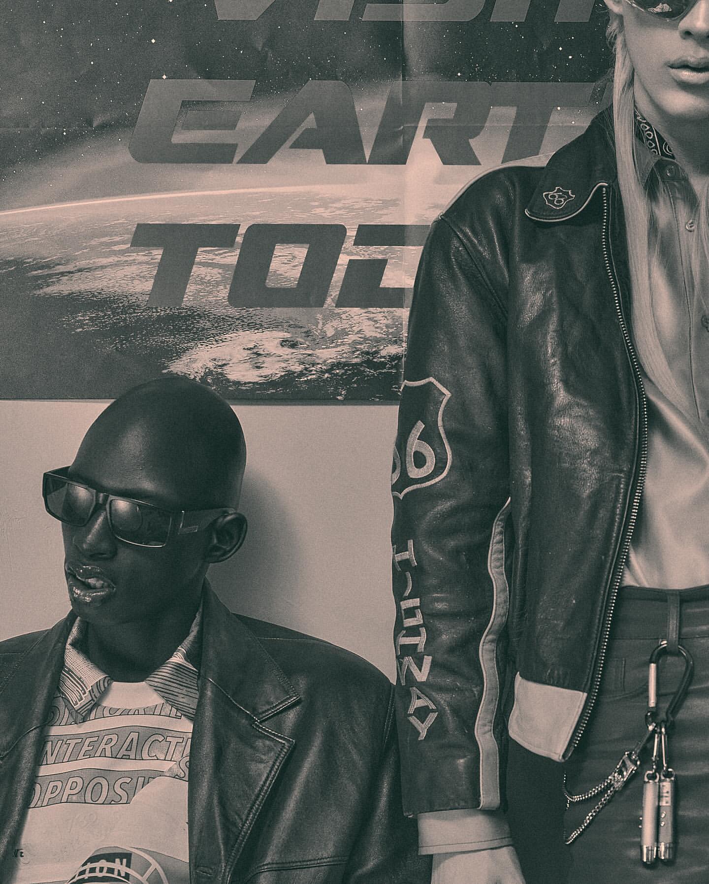 Two adult fashion models pose indoors against a wall with a poster of Earth. One model is seated wearing sunglasses and a leather jacket.