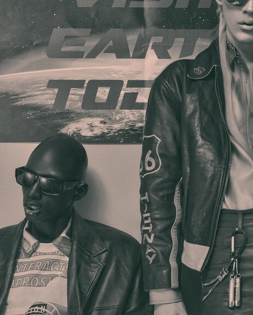 Two adult fashion models pose indoors against a wall with a poster of Earth. One model is seated wearing sunglasses and a leather jacket.