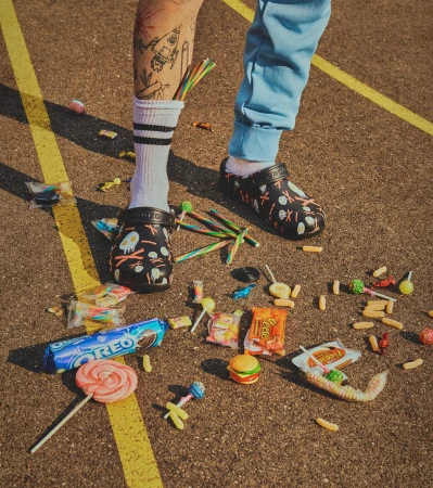 Person wearing Crocs with cartoon pattern surrounded by colorful candy and packaged sweets on paved ground with yellow lines.