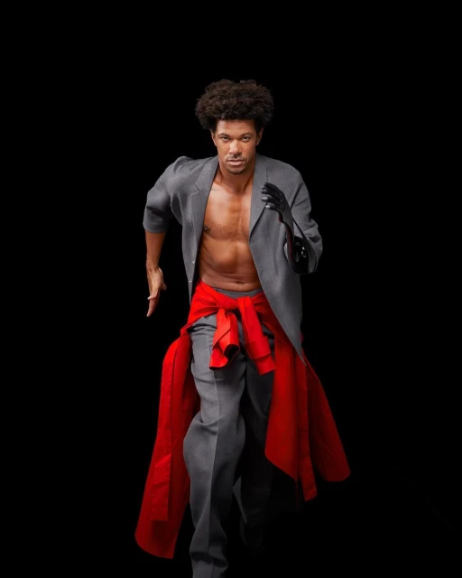 Man in a grey suit and red sash runs toward the camera in a studio shot with a black background, demonstrating athletic fashion.