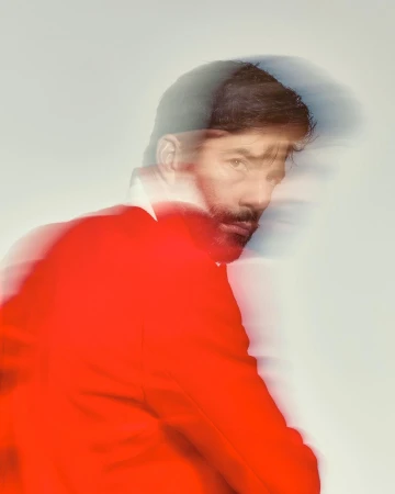Portrait of a bearded man wearing a vibrant red jacket with motion blur effect against a plain white studio background.