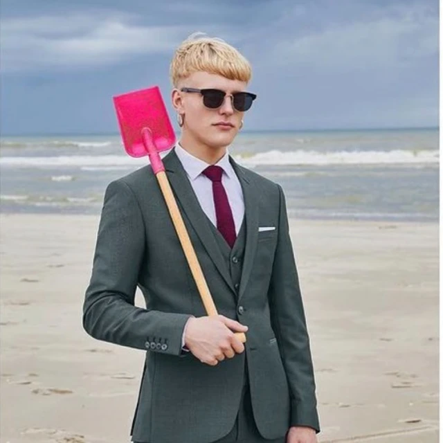 Young man in a suit stands on a sandy beach carrying a pink toy shovel, with the ocean and sky in the background on a cloudy day.
