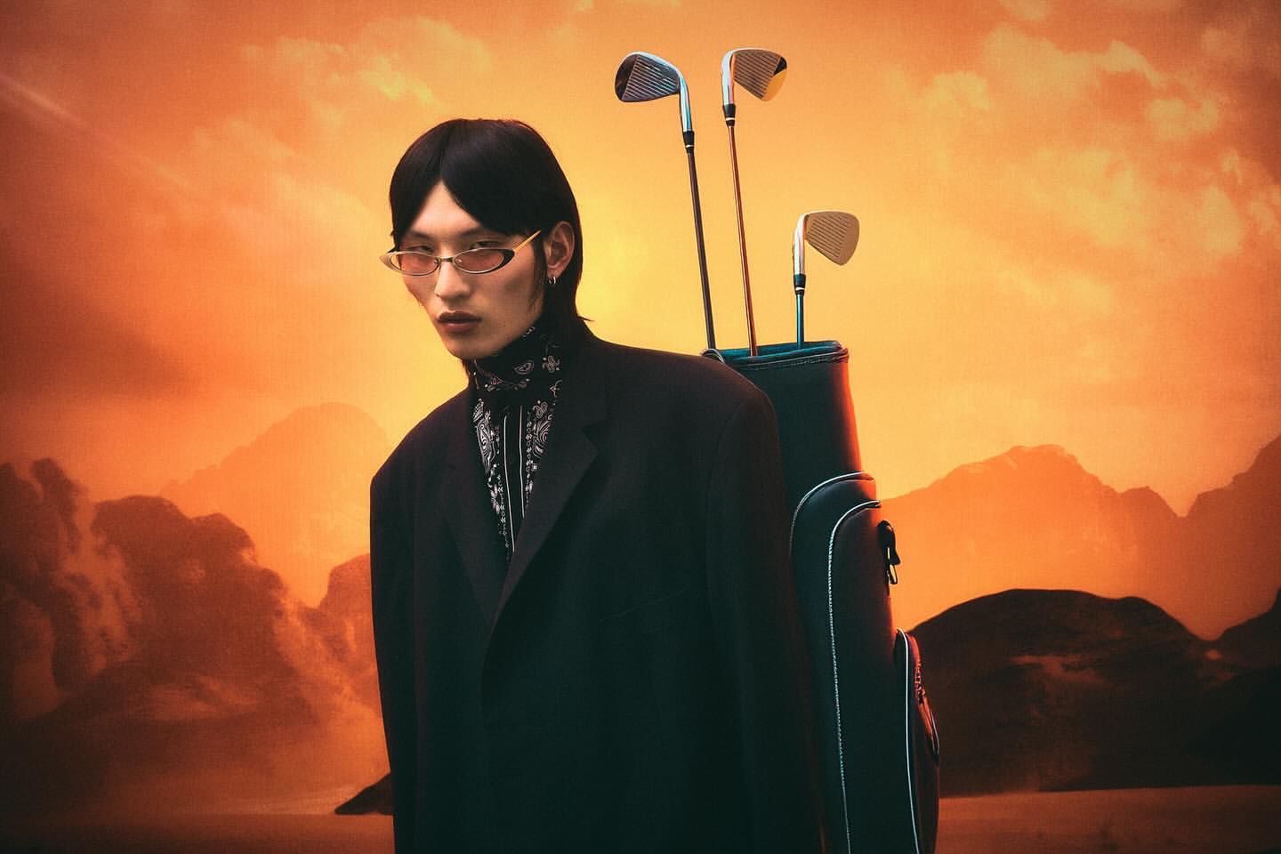 Stylish Asian man in a black suit and retro glasses poses with a golf bag and clubs against an orange, hazy mountain backdrop.