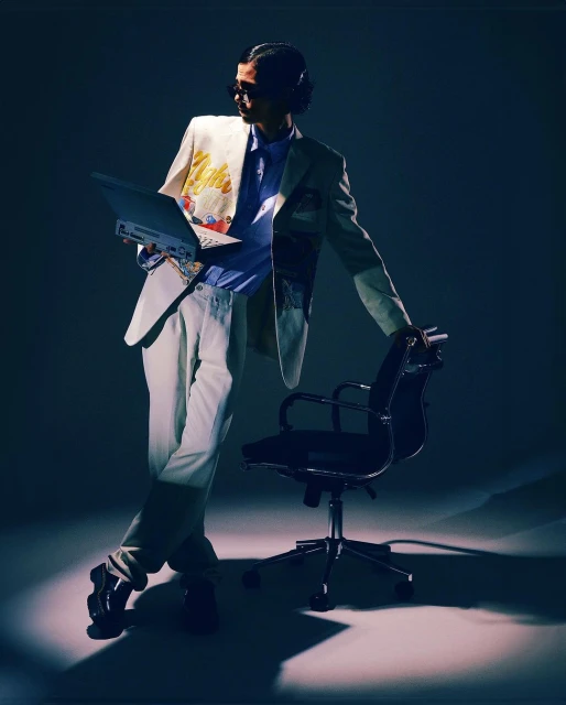 Man in light suit poses with laptop and chair in dramatic lighting, wearing headphones and sunglasses for modern, stylish portrait.