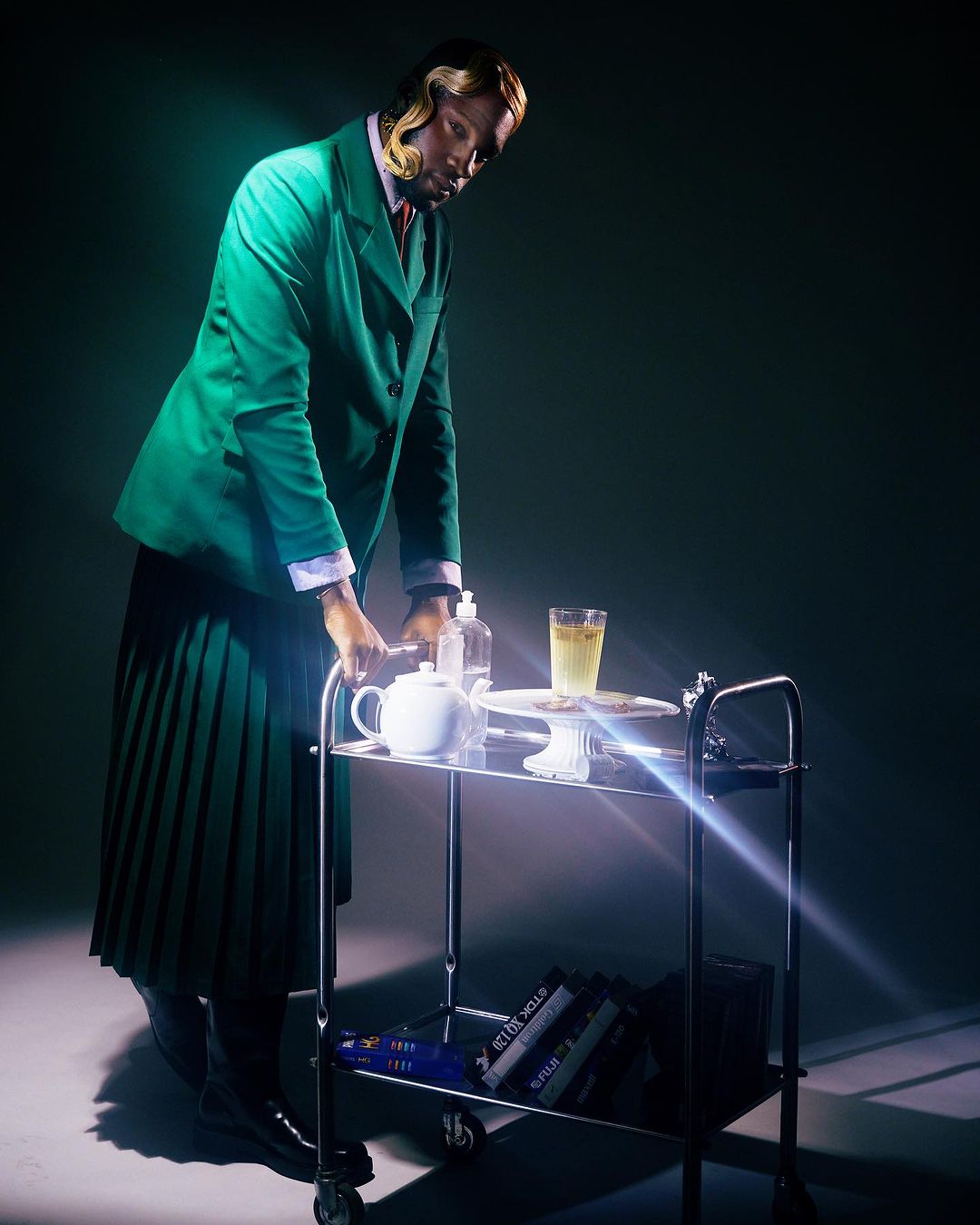Stylish person in green suit and pleated skirt leans on a silver tea cart with tea set and books in a dark, dramatic studio shot.