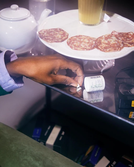 Adult hand reaches for small, white pills on a reflective surface near a white teapot, sliced meat, and a glass of yellow liquid.