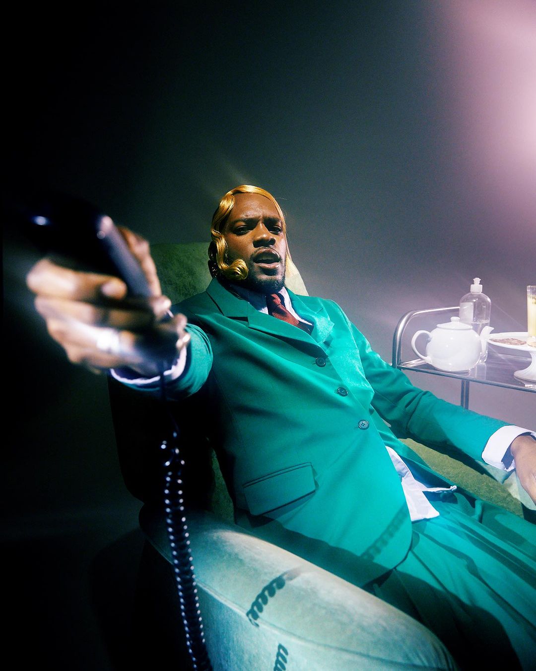 Man in green suit holds telephone receiver in a studio. He has a blond wavy wig, and there is a table with a tea set nearby.