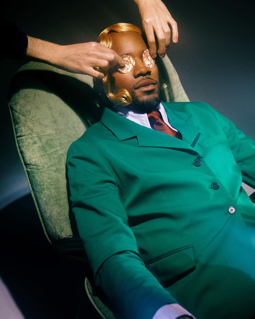 Man in green suit reclines in chair with blonde wig, eyes covered with sliced sausage by disembodied hand in dramatic, moody lighting.