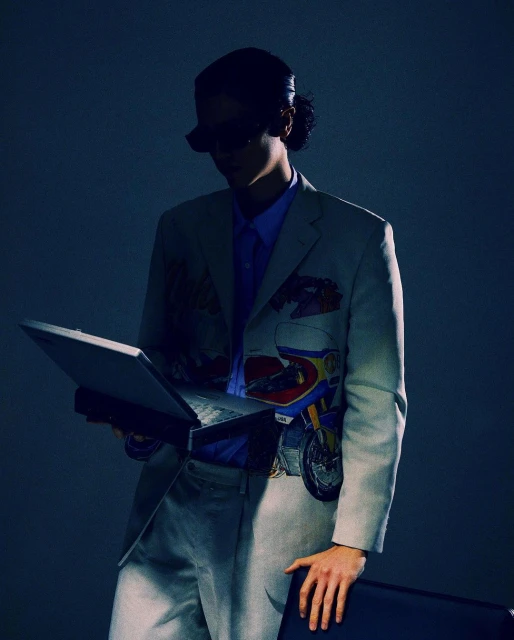 Adult poses with a laptop in a stylish cream-colored suit jacket with a motorcycle graphic in an indoor studio with cool lighting.