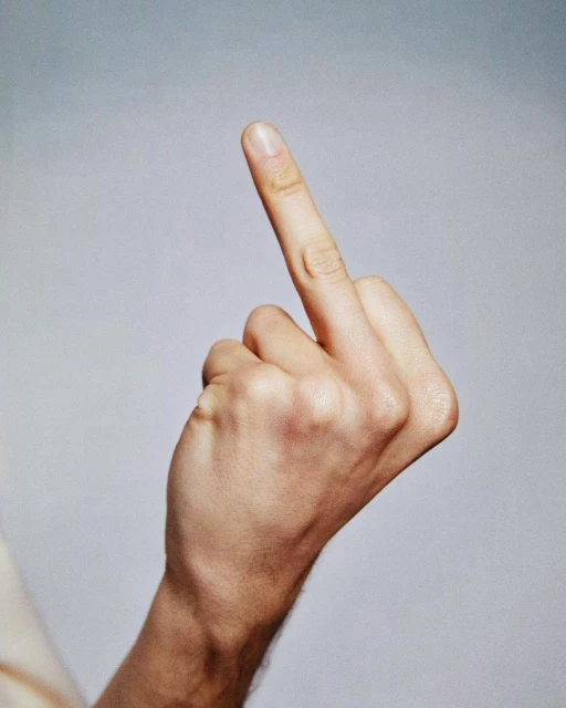 Close-up of a light-skinned human hand gesturing with the middle finger extended against a neutral gradient background in a studio setting.