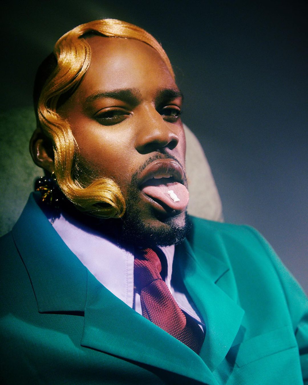 Dark-skinned adult man with a pill on his tongue, light brown vintage-styled hair, and a green suit, posing indoors.