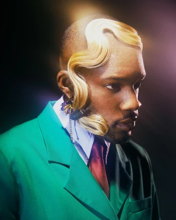 Stylish man in a green suit with unique blonde hair, accessorized with purple and black earrings, set against a dark background.