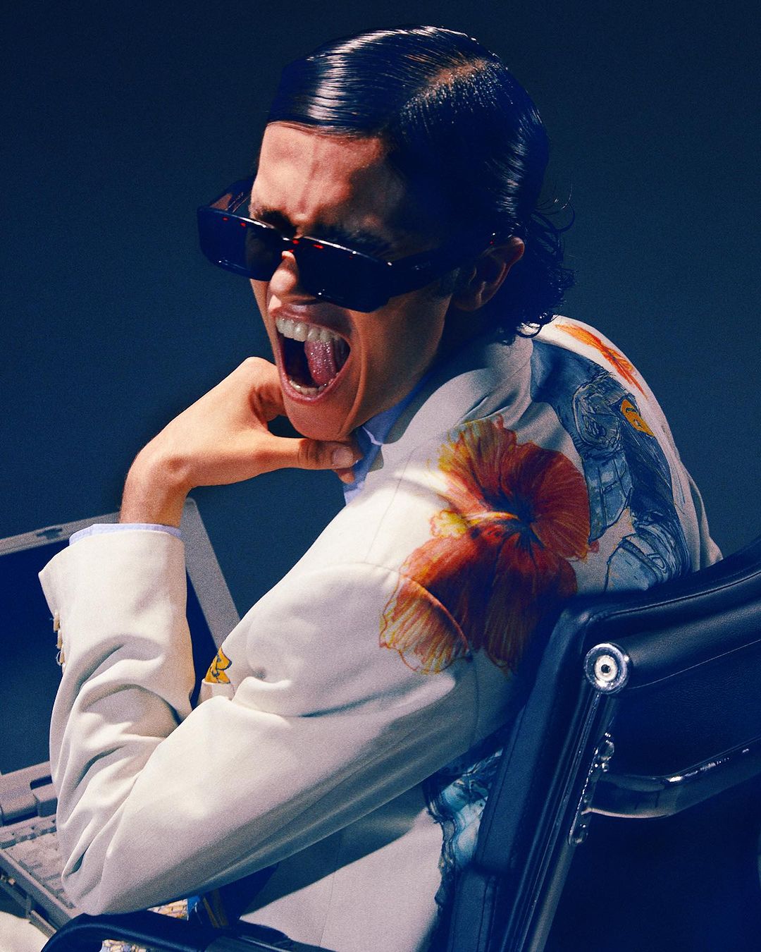 Person with slicked-back dark hair and sunglasses, wearing a floral jacket, sitting in an office chair and yelling with their mouth wide open.
