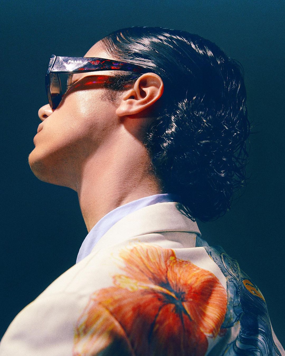 Stylish portrait of a person wearing sunglasses and a floral jacket, shot from a low angle against a dark blue background.