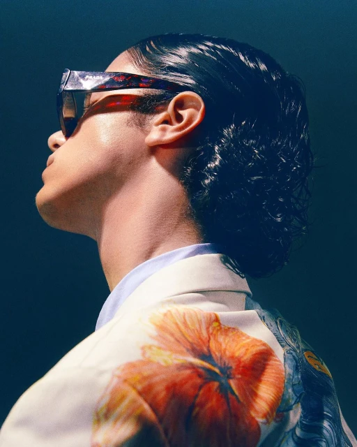 Stylish portrait of a person wearing sunglasses and a floral jacket, shot from a low angle against a dark blue background.