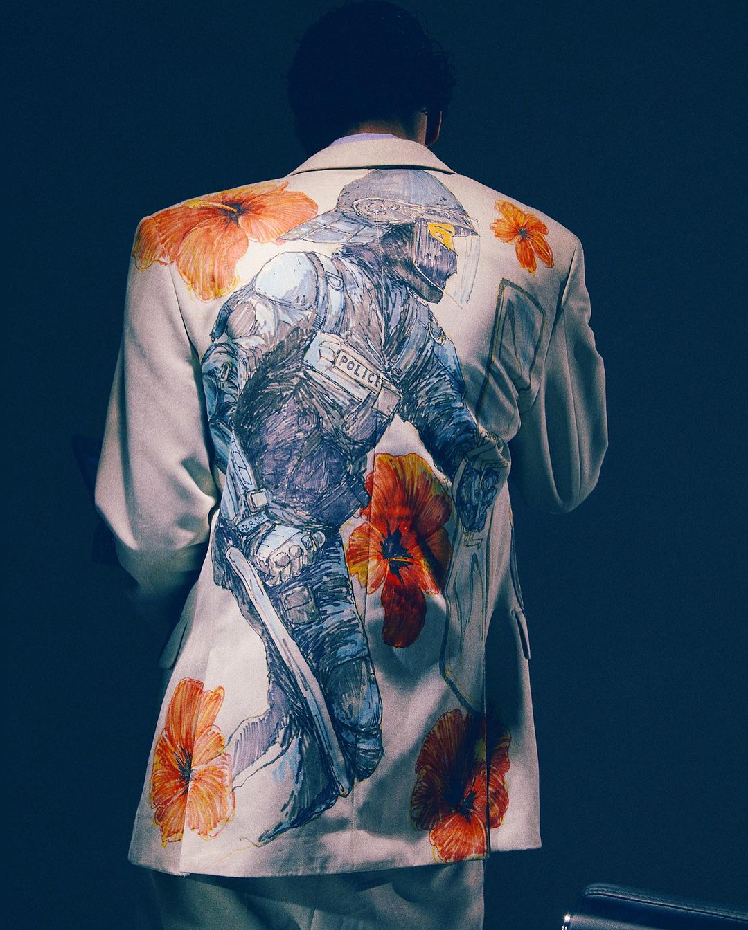 Back view of a person wearing a white suit jacket with a drawing of a police officer and orange flowers against a dark blue background.