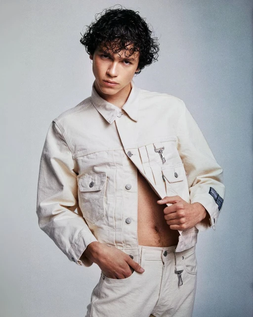 Man poses in off-white denim jacket and pants, showcasing a modern, casual style against a neutral backdrop in a fashion editorial.