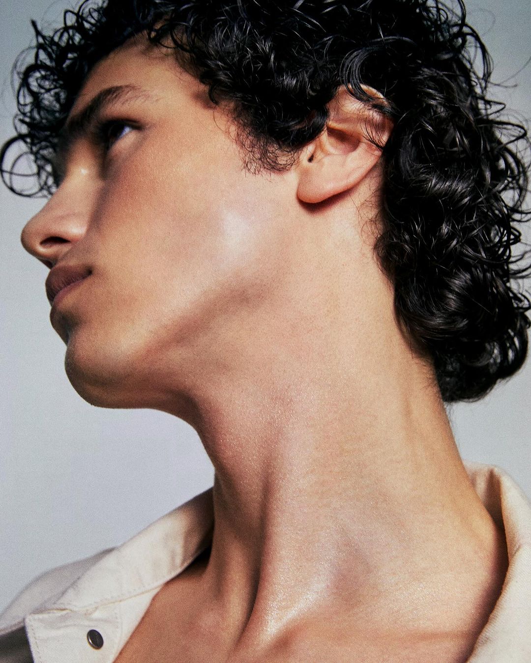 Close-up studio shot of a male model with dark, curly hair and tan skin, gazing upward. He's wearing a collared shirt against a neutral background.