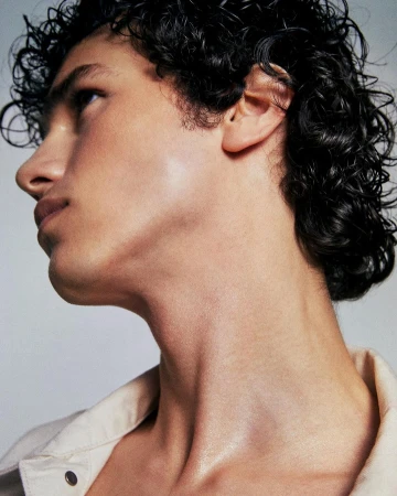 Close-up studio shot of a male model with dark, curly hair and tan skin, gazing upward. He's wearing a collared shirt against a neutral background.