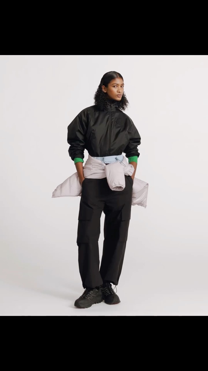 Woman in a black nylon jacket and black cargo pants stands in a bright studio, with a sweater tied around her waist. Fashion.
