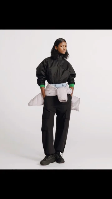 Woman in a black nylon jacket and black cargo pants stands in a bright studio, with a sweater tied around her waist. Fashion.