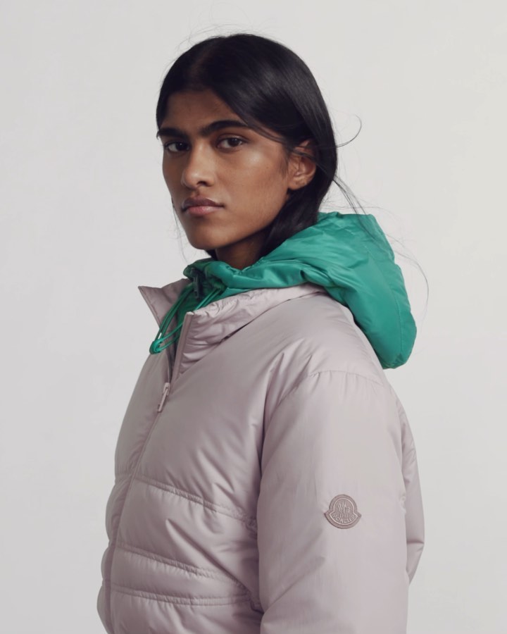 A woman poses in a lavender puffer jacket with a green hood against a white background in a studio shot, looking at the camera.