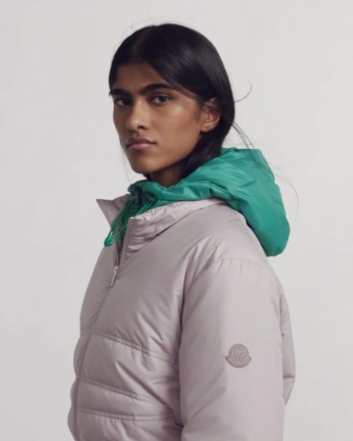 A woman poses in a lavender puffer jacket with a green hood against a white background in a studio shot, looking at the camera.