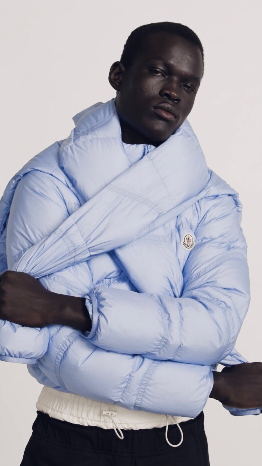 Fashionable African-descent adult model poses in a light-blue puffer jacket against a plain background in a studio setting. Minimalist and clean.