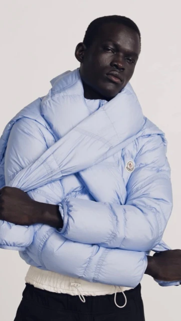 Fashionable African-descent adult model poses in a light-blue puffer jacket against a plain background in a studio setting. Minimalist and clean.