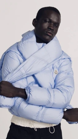 Fashionable African-descent adult model poses in a light-blue puffer jacket against a plain background in a studio setting. Minimalist and clean.