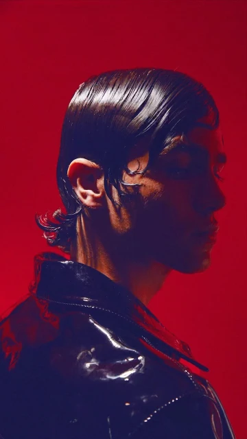 Man in profile wearing a black patent leather jacket against a red background, showcasing studio lighting and fashion style.