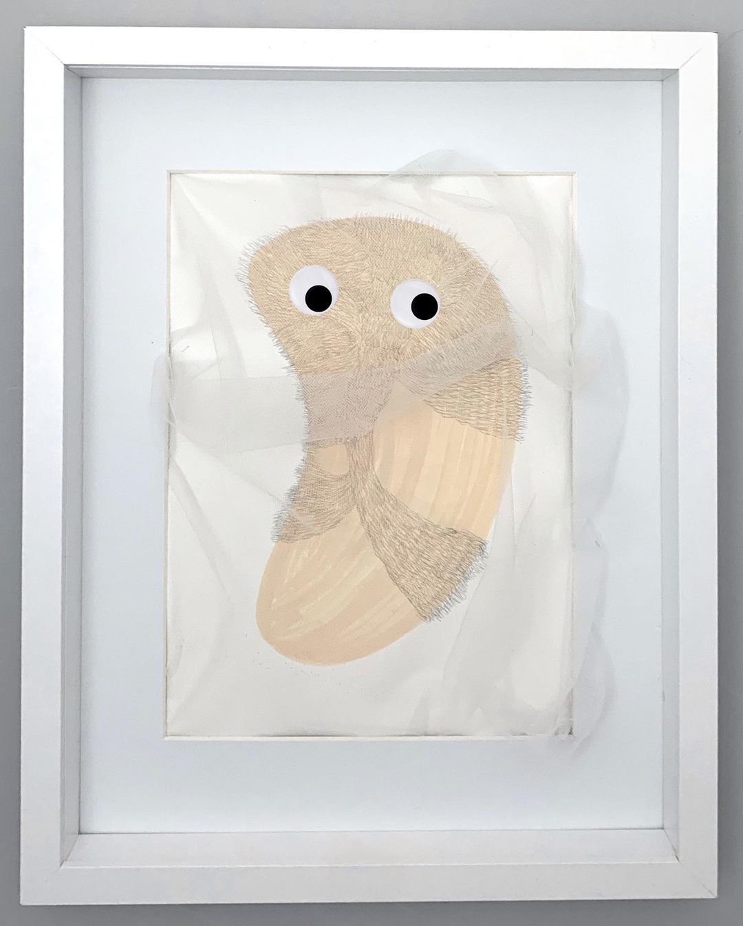 Framed art features a textured, tan abstract form with googly eyes on a white background, encased in a white frame against a grey wall.