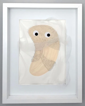 Framed art features a textured, tan abstract form with googly eyes on a white background, encased in a white frame against a grey wall.