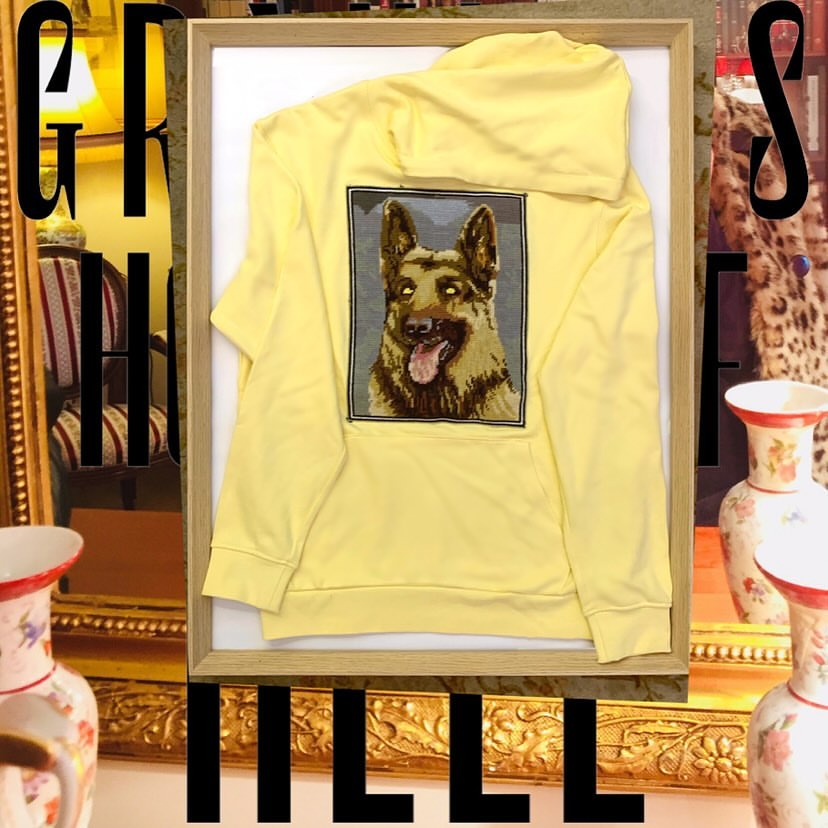 A yellow hooded sweatshirt with an embroidered German Shepherd on the back, presented in a light wood frame against a still life backdrop.