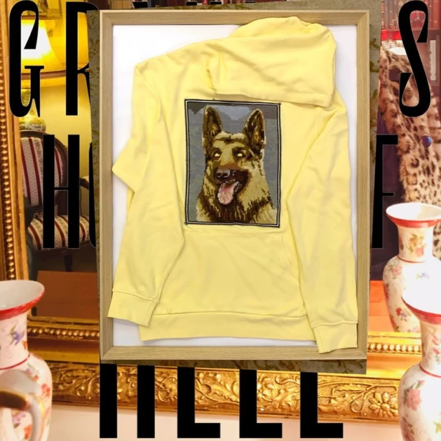 A yellow hooded sweatshirt with an embroidered German Shepherd on the back, presented in a light wood frame against a still life backdrop.