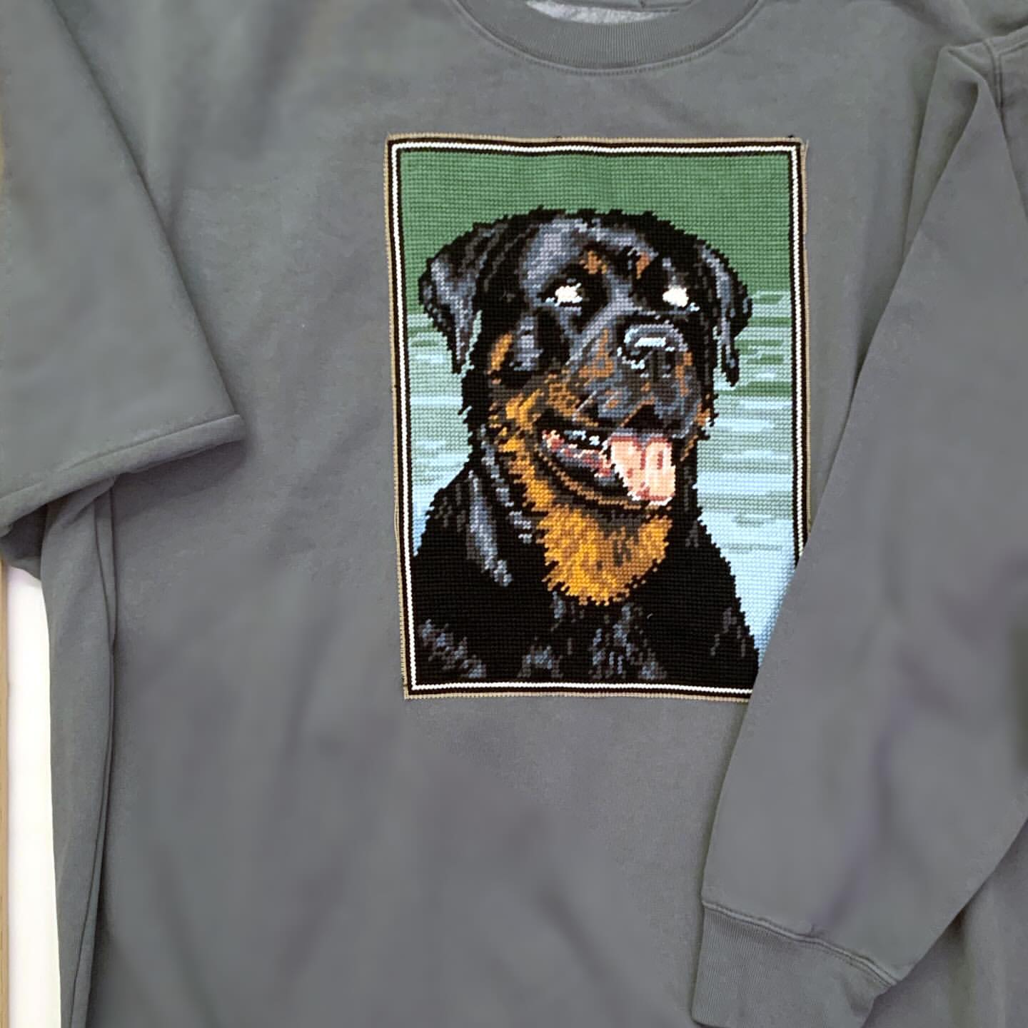 Grey sweatshirt featuring a needlepoint-style Rottweiler dog design. The design is framed and shows the dog's head and shoulders.