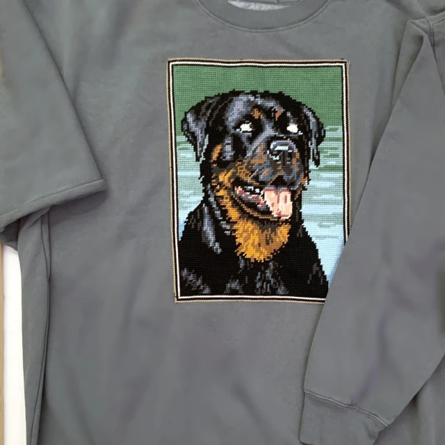 Grey sweatshirt featuring a needlepoint-style Rottweiler dog design. The design is framed and shows the dog's head and shoulders.